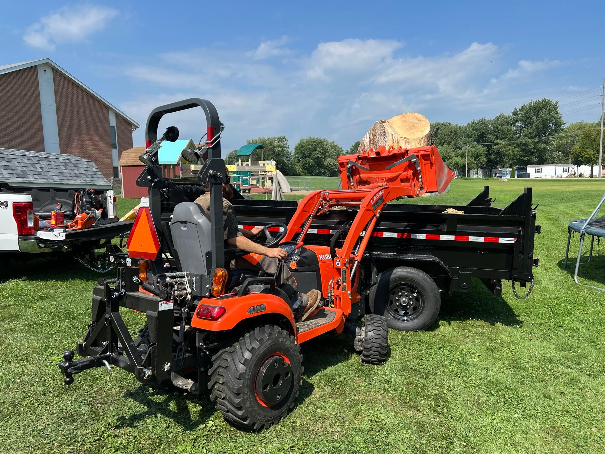 Compact tractor with a loader arm lifting a large cut log into a dump trailer