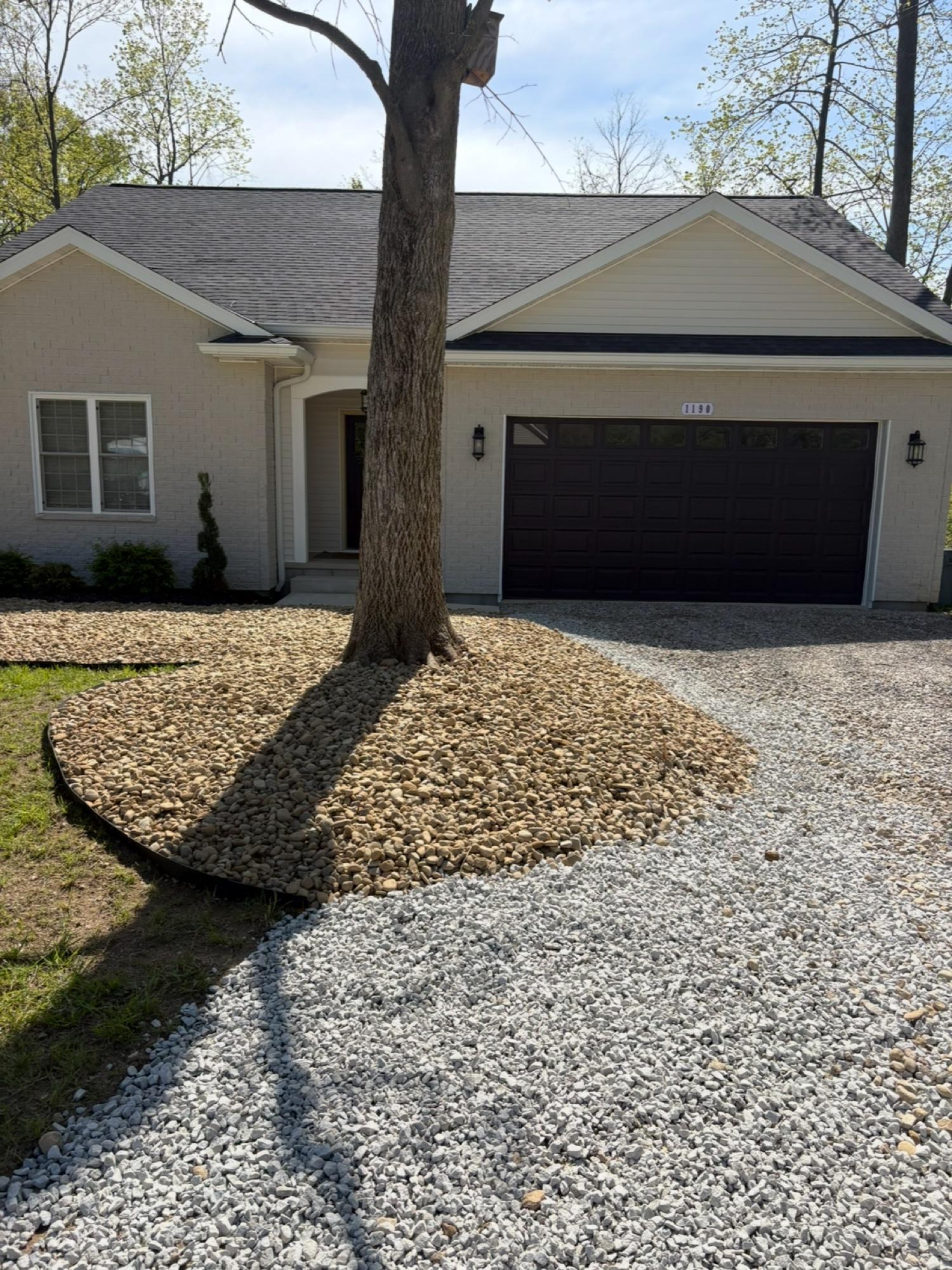 Finished river rock landscaping bed around a front-yard tree with a matching gravel driveway